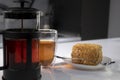 cup of tea in a double-botoom glass, french-press teapot and piece of cake on white table in gray kitchen