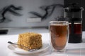 cup of tea in a double-botoom glass, french-press teapot and piece of cake on white table in gray kitchen