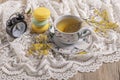 A cup of tea next to an alarm clock that strikes five o\'clock on a lace tablecloth with three stacked macarons