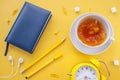 Cup of tea with splashes, lumps of sugar, notebook in blue cover, pen and pencil, yellow alarm clock,