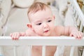 A curious baby is standing in a crib, holding the handrails and smiling. Funny lop-eared child in bed, six months old