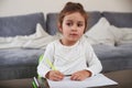 A curious beautiful little girl sitting at a table holds a pen and looks away. Drawing at home Royalty Free Stock Photo