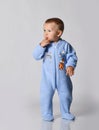 Blond baby boy toddler in blue cotton jumpsuit with rainbow and tiger stands holding finger at mouth and looks aside