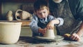 Curious boy is learning pottery from his experienced grandfather in small home studio. Child is forming clay to make Royalty Free Stock Photo