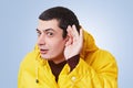 Curious brunet male model tries to overhear something, keeps hand on ear, listens gossips in next room, wears yellow anorak, isola