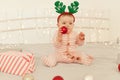 Curious cute infant girl sitting on bed and wearing striped long sleeve baby sleeper and festive deer horns, biting ball for Royalty Free Stock Photo