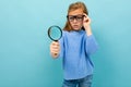 Curious european girl in glasses looks through a magnifying glass on a light blue background