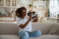 Curious excited young African woman holding modern VR glasses while sitting on sofa in living room