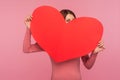Curious funny woman in pink sweater taking look hiding behind big paper heart, saint valentine day celebration