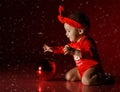 Infant baby girl toddler in red bodysuit and plays with catches Christmas New Year fir tree decoration ball under snow Royalty Free Stock Photo