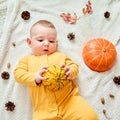 A curious infant baby is lying on a blanket in yellow autumn clothes with a red pumpkin. Concerned child with orange leaves, top