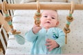 Curious infant baby is playing with wooden hanging toys lying in bed. Happy funny child in turquoise clothes in a crib, aged six