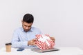 Curious man employee sitting in office workplace, opening gift box and looking inside, unwrapping present