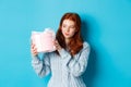 Curious teen girl with red hair, shaking gift box and wonder what inside, standing over blue background