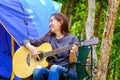 Curly blond long hair asian teenager girl with long shrit and white shorts playing guitar front of blue canvas camping tent on Royalty Free Stock Photo