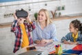 Curly boy and dark-haired girl sitting at round table with blonde female, boy wearing vr glasses, feeling excited Royalty Free Stock Photo