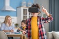 Curly boy wearing vr glasses, looking excited, dark-haired girl and blonde woman sitting at round table, busy with tasks Royalty Free Stock Photo