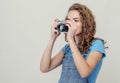 Curly brunette girl in denim overalls is holding a vintage film camera in her hands. Takes a picture. Side view