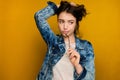 A curly dark-haired girl in a jeans stands on a yellow background, looks to the side, pressing the earpiece of glasses