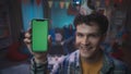 Curly guy in a festive hat, smiling, shows a phone with a green screen. Close up of a guy with a smartphone on a blurred Royalty Free Stock Photo