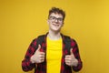 Curly guy student in glasses with a backpack stands on a yellow background and shows likes with his hands
