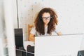 Curly hair girl teenager in glasses and headphones in white t-shirt sitting working on computer at bright room at home