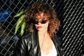 Curly hair woman in sunglasses and leather jacket posing on grid background