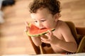 Curly-haired baby girl eats fruit sitting indoors at home in summer