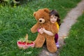 A curly-haired girl in a yellow T-shirt has planted a big brown teddy bear on her knees and is squatting. near a basket with Royalty Free Stock Photo