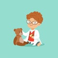 Curly-haired toddler boy examining his patient teddy bear toy. Baby character in white doctor s coat, pants, shirt and