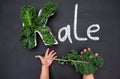 Curly-leaf kale cabbage inside letter K shaped plate, chalk inscription KALE on blackboard, child`s hands holding fresh greens.