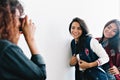 Curly tanned female photographer with red manicure taking photo of glad latin and asian girls in sport jackets. Adorable