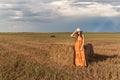 Curly young rural teenager girl stands near a bundle of hay in a sundress and hat on a harvested wheat field with a backdrop of a Royalty Free Stock Photo
