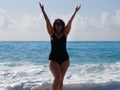 A curvaceous woman in a black bathing suit stands in the sea, raising her hands