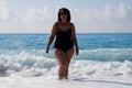 A curvaceous woman in a black bathing suit and in sunglasses stands in the sea, raising her hands