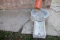 Curved end of brown drainpipe at corner of house with chipped plaster wall and cracked cement blind area. Under pipe concrete tray