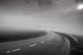 Curving, wet asphalt road in dense fog, sun shining behind thick clouds,