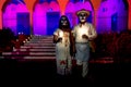costumed man and woman with with mexican traditional white clothes and skull make-up in front of red pink lightened building with