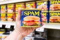 Customer hand holding a tin can of Spam Brand meat in a supermarket aisle