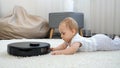 Cut elittle baby lying on carpet at house and looking at robot vacuum cleaner doing cleanup on carpet Royalty Free Stock Photo