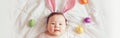 Cute adorable Asian baby wearing pink Easter bunny ears. Infant kid lying on bed with coloredl Easter eggs. Funny child