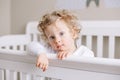 Cute adorable baby boy toddler standing in crib at kids nursery room at home. Curious charming baby boy with curly blonde hair and