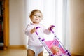 Cute adorable baby girl making first steps with doll carriage. Beautiful toddler child pushing stroller with toy at home Royalty Free Stock Photo