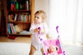 Cute adorable baby girl making first steps with doll carriage. Beautiful toddler child pushing stroller with toy at home Royalty Free Stock Photo