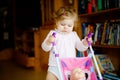 Cute adorable baby girl making first steps with doll carriage. Beautiful toddler child pushing stroller with toy at home Royalty Free Stock Photo