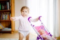 Cute adorable baby girl making first steps with doll carriage. Beautiful toddler child pushing stroller with toy at home Royalty Free Stock Photo