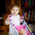 Cute adorable baby girl making first steps with doll carriage. Beautiful toddler child pushing stroller with toy at home Royalty Free Stock Photo