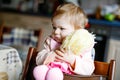 Cute adorable baby girl playing with first doll. Beautiful toddler child sitting in high chair at home. Happy healthy