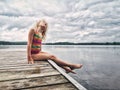 Cute adorable blonde Caucasian girl sitting on wooden dock by lake and looking up in the sky. Dreaming relaxing cute child kid by Royalty Free Stock Photo