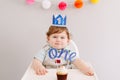 Cute adorable Caucasian baby boy in blue crown celebrating first birthday at home. Child kid toddler sitting in high chair eating Royalty Free Stock Photo